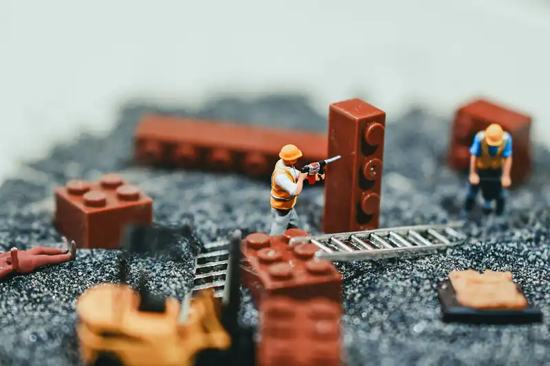 Tiny model builders in hard hats work among brown toy bricks on grey gravel